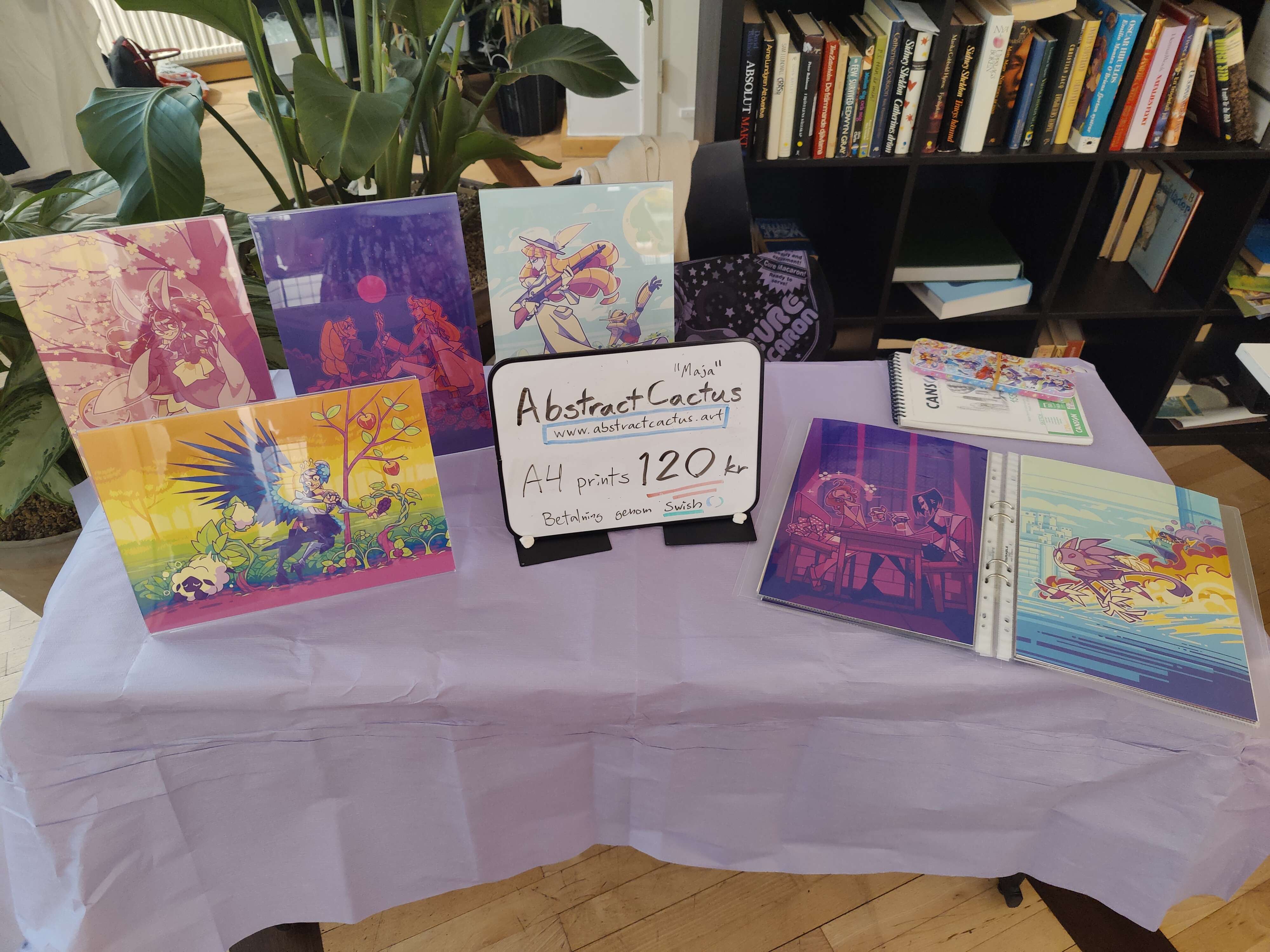A photo of my artist alley setup, with 4 pictures displayed in acrylic stands and a binder for browsing.