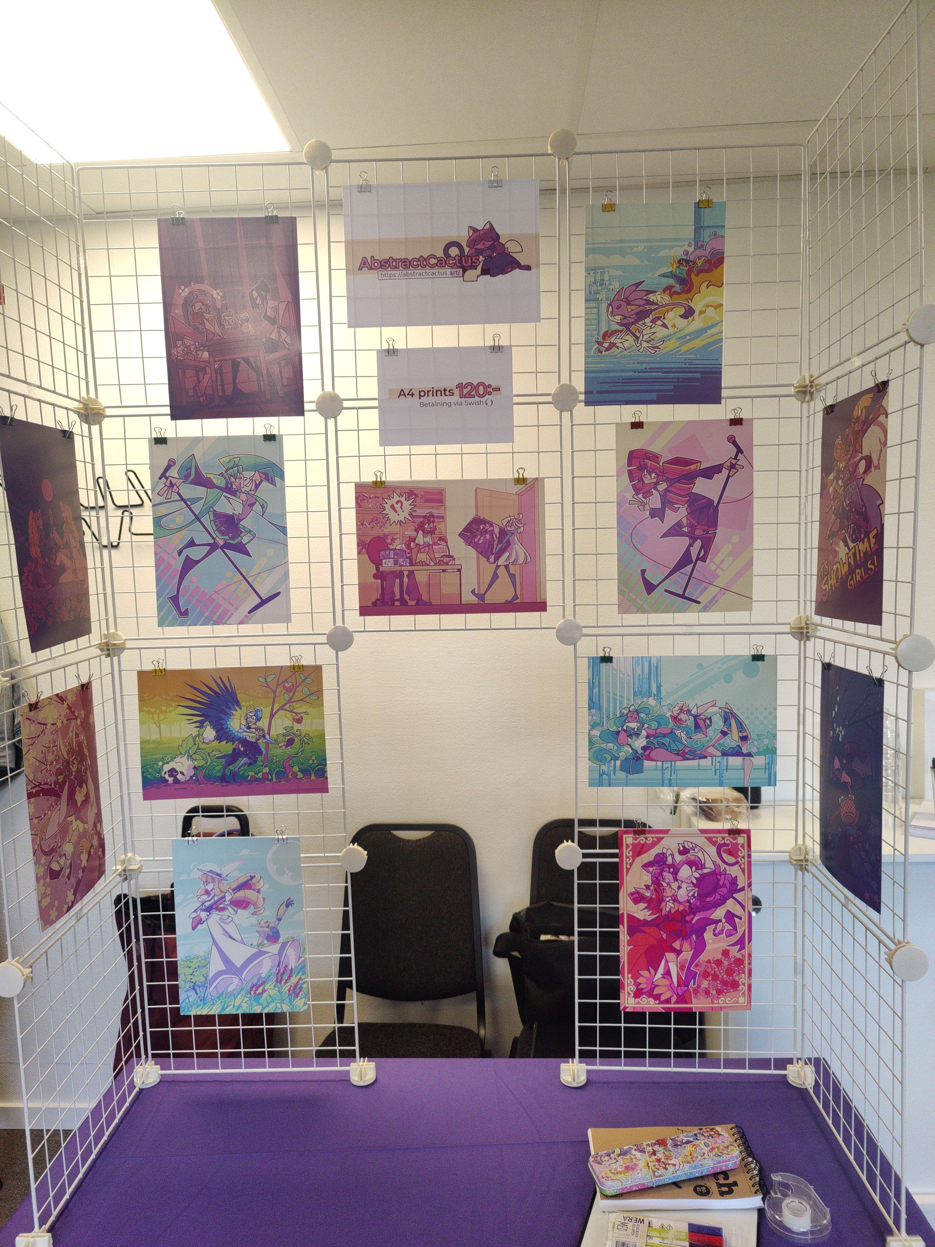 A photograph of my shoddy Artist Alley table, with a white wire grid display and a bunch of A4 art prints hanging on it.