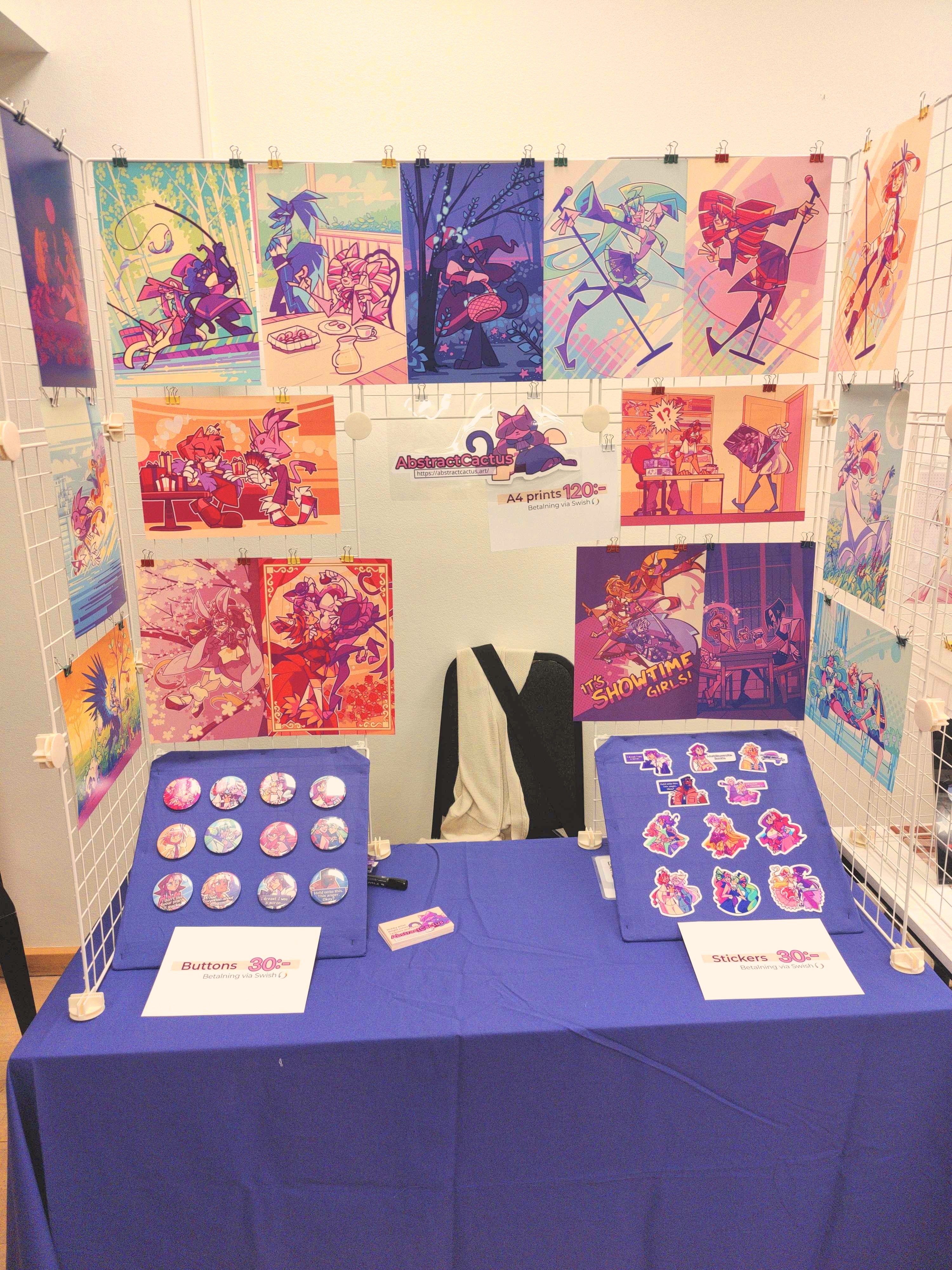 A photograph of my not as shoddy Artist Alley table, a white wire grid display with a bunch of A4 art prints hanging on it and two panels down on the table with pins and stickers on them.